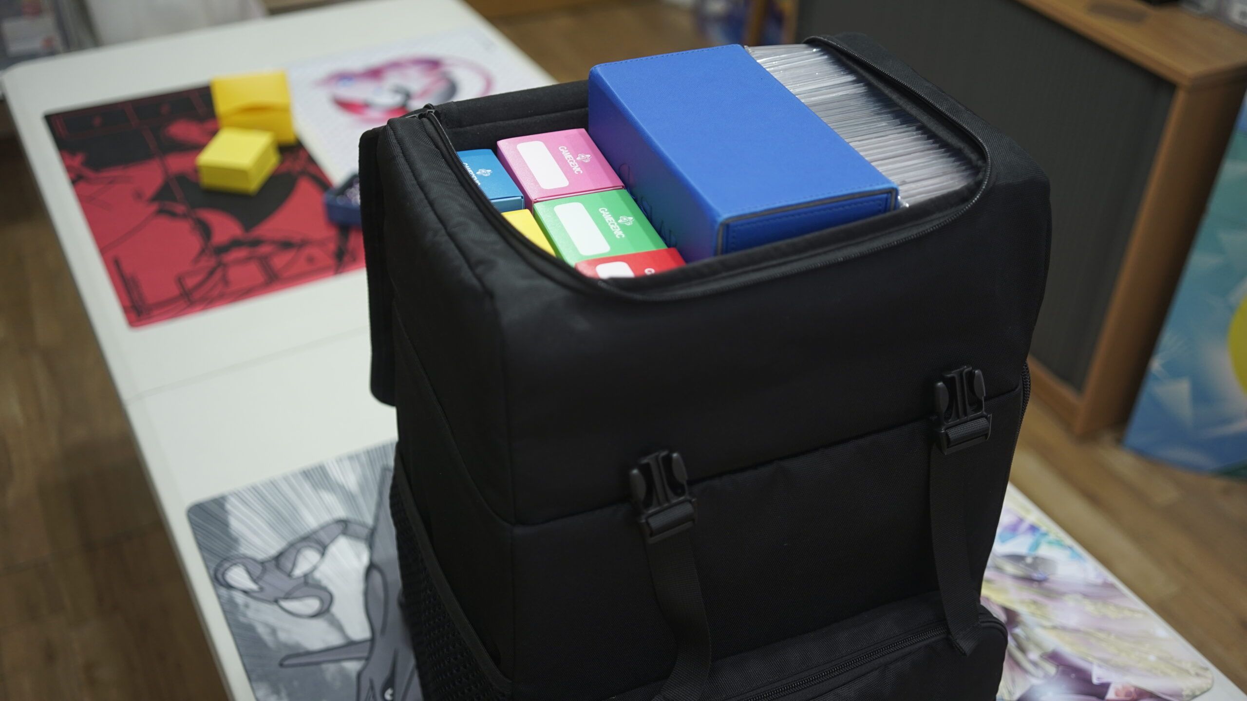 QUAD TCG backpack on a game table at a TCG league night shown holding deck boxes and graded cards in the top compartment
